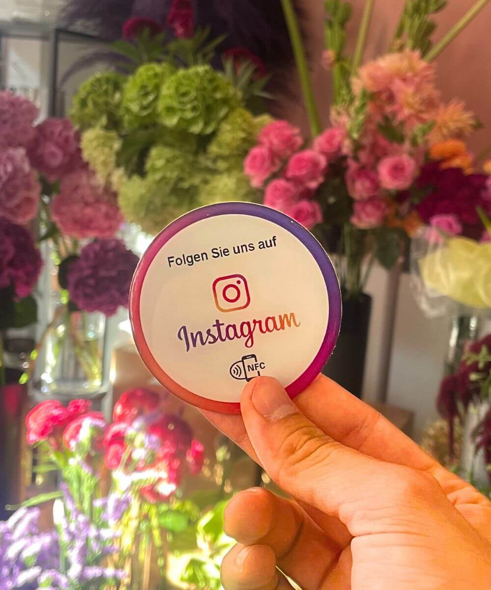 Ein NFC Sticker mit der Aufschrift "Folgen Sie uns auf Instagram". Die BLumen im Hintergrund haben die selbe Farbe wie der Instagram Sticker. Der Instagrma Sticker hat die Farben von Instagram. Innen weiß.